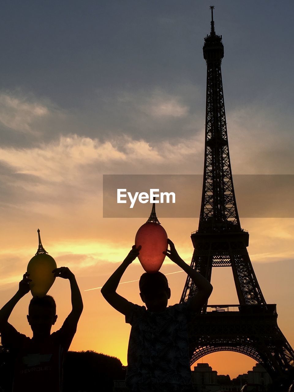Low angle view of silhouette sibling with balloons near eiffel tower against sky during sunset