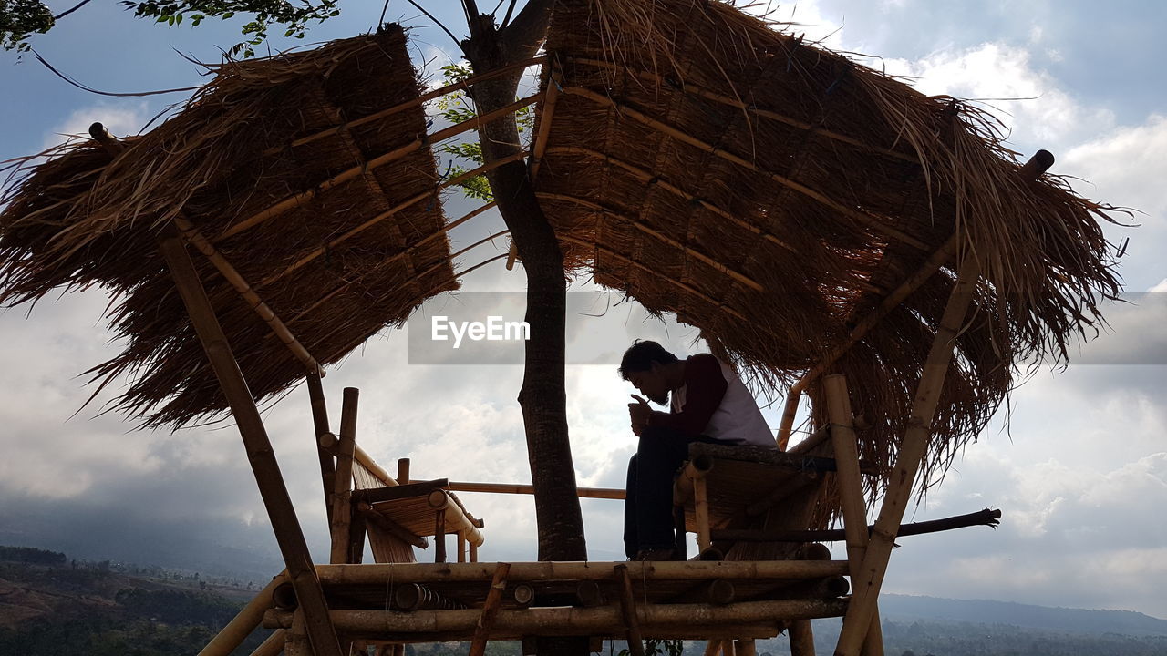 Man smoking cigarette while sitting in hut against sky