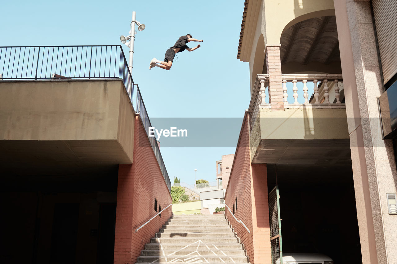 From below side view of strong male doing parkour and jumping from metal railing on brick building while showing stunts in city