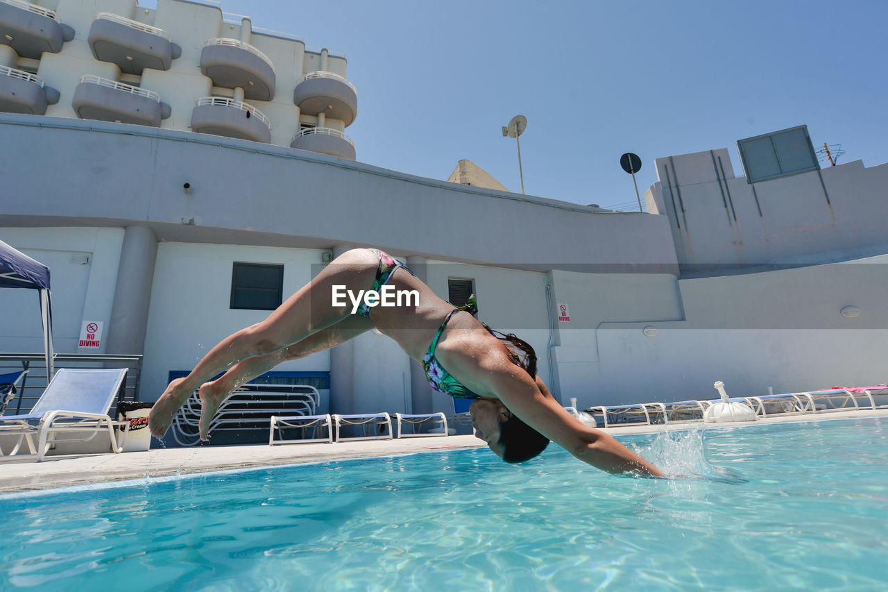 LOW ANGLE VIEW OF MAN JUMPING IN SWIMMING POOL AGAINST SKY