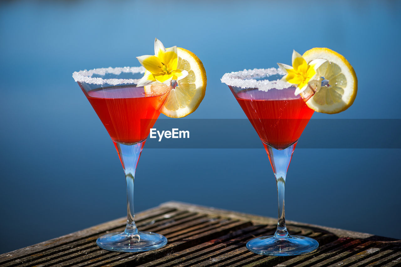 Drink garnished with yellow flower with lemon slice on wooden table against swimming pool