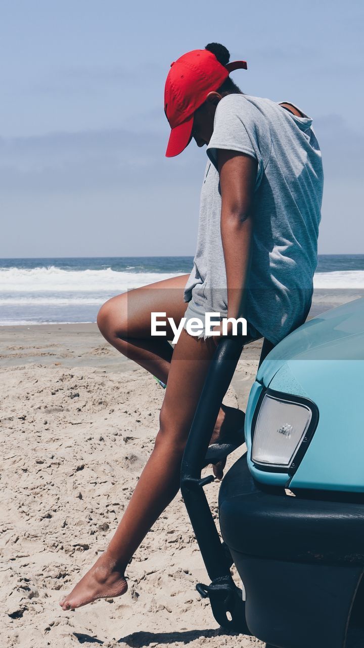 Side view of woman sitting on car at beach against sky