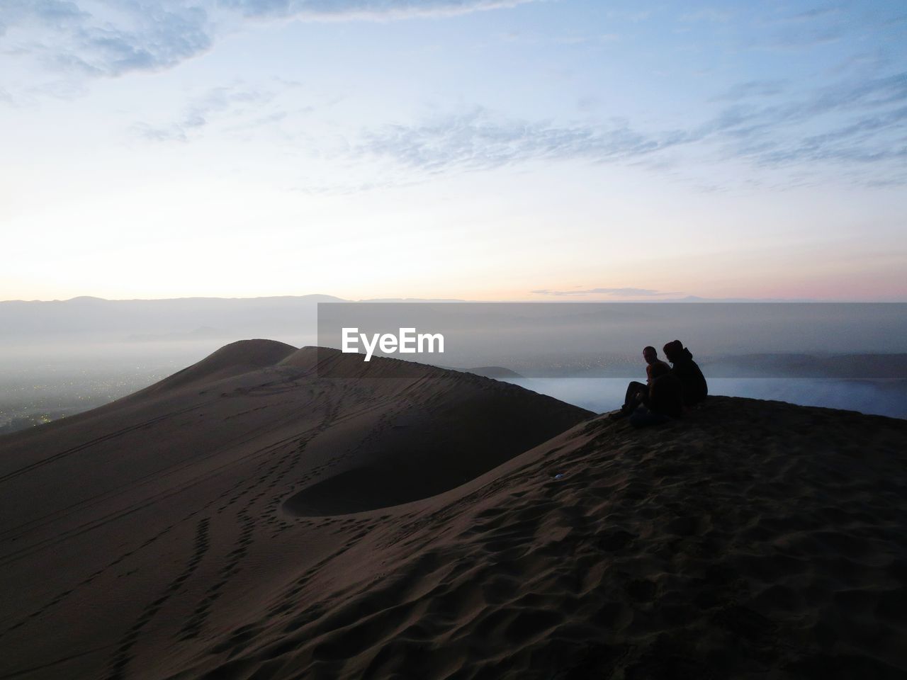 Friends sitting on sand dune against sky during sunset