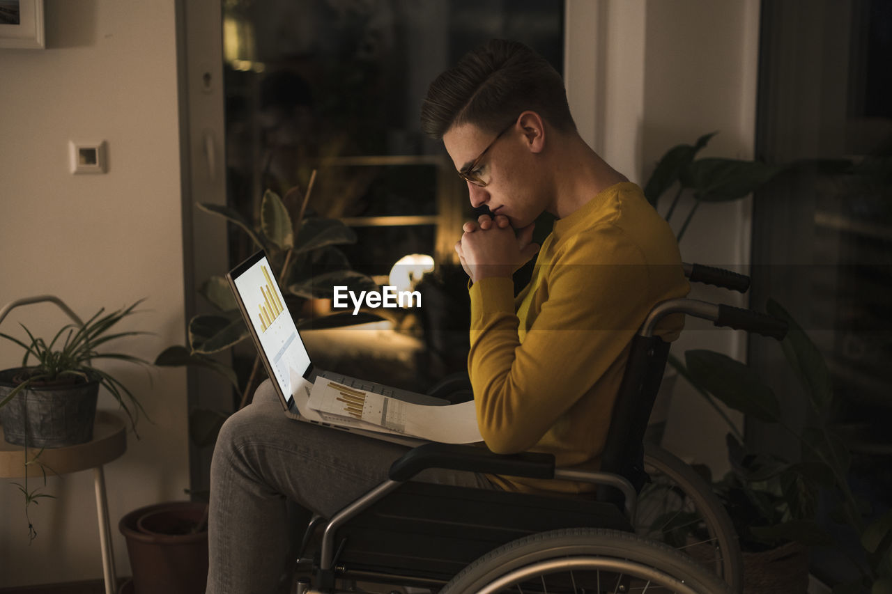 Concentrated oung man in wheelchair with laptop working late while sitting on wheelchair in living room