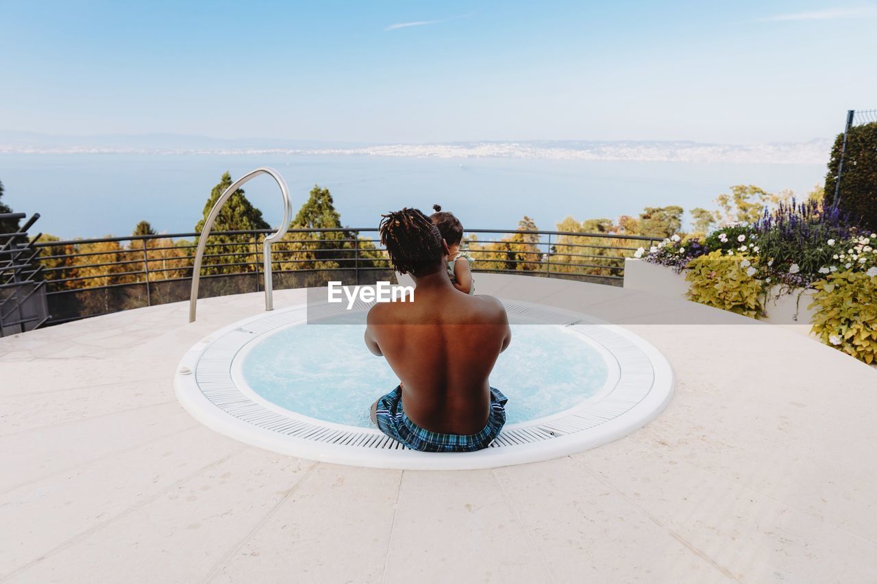 Rear view of father holding daughter while sitting in swimming pool against sea and sky