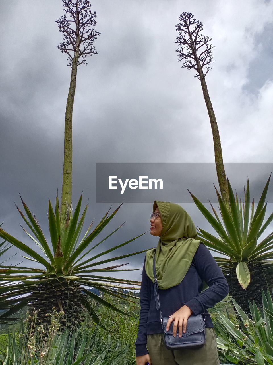 Woman wearing hijab standing amidst plants against cloudy sky