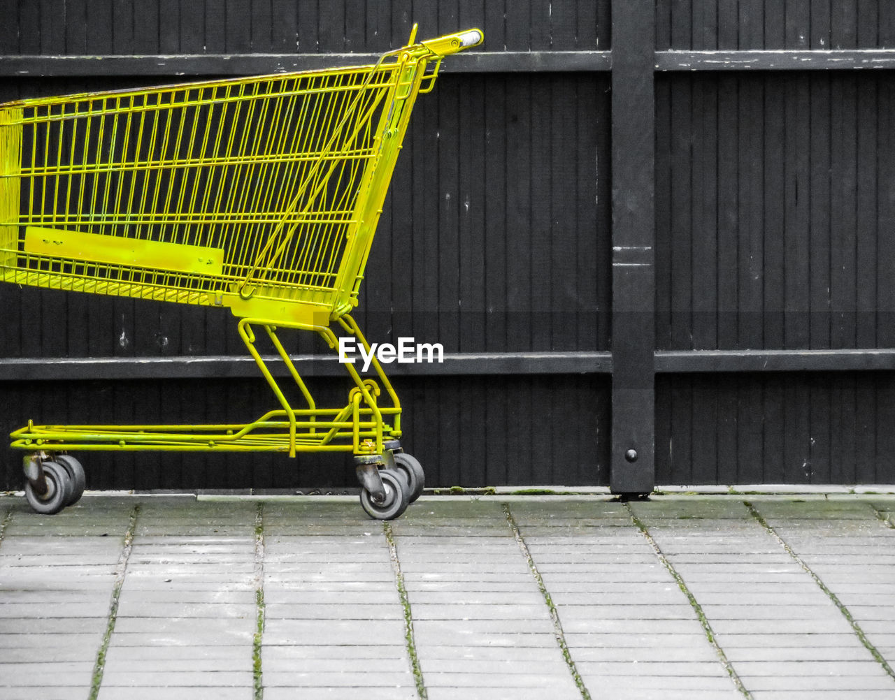 Empty yellow shopping cart on footpath against fence