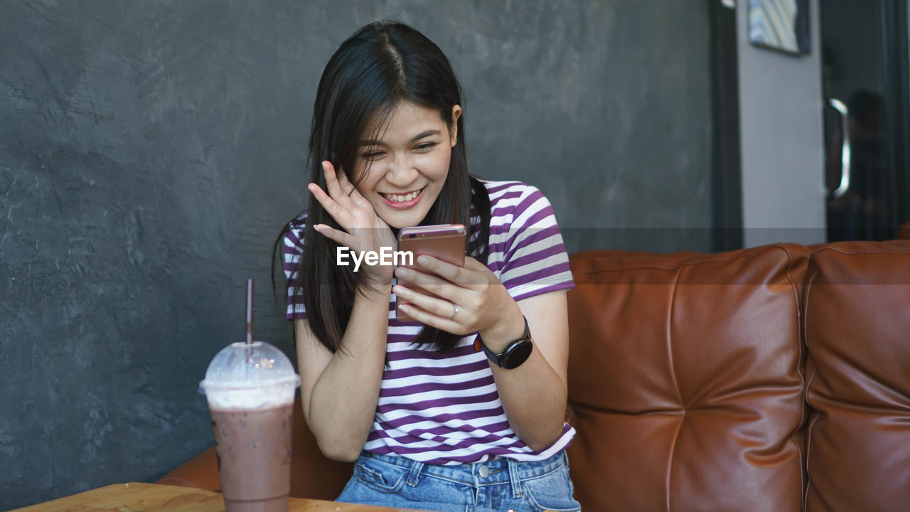 Cheerful young woman using smart phone sitting at table with coffee in cafe