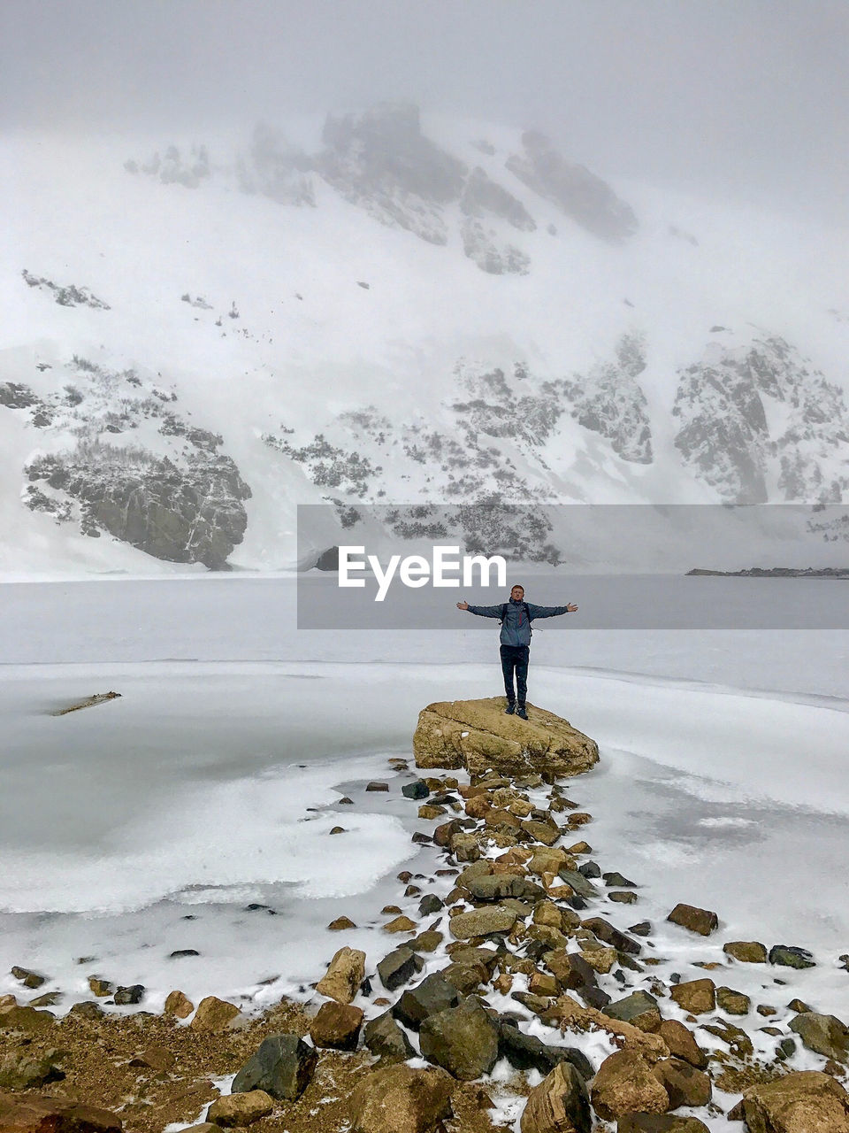 Man with arms outstretched standing on rock against lake during winter