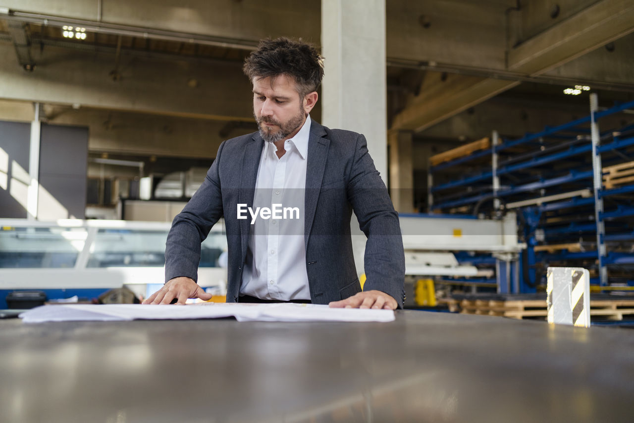 Mature businessman in suit working while standing by table at factory
