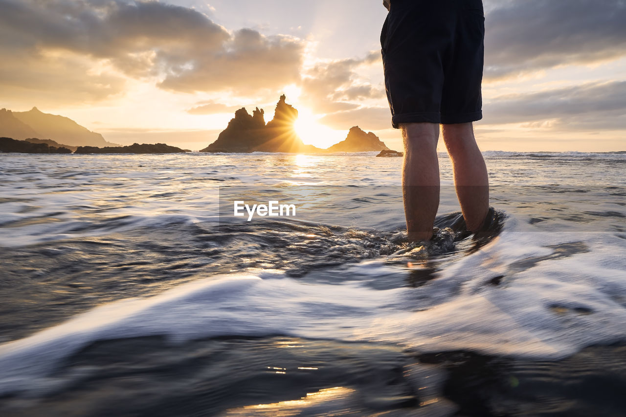 Low section of man during relaxation in ocean. benijo beach against sky at beautiful sunset.