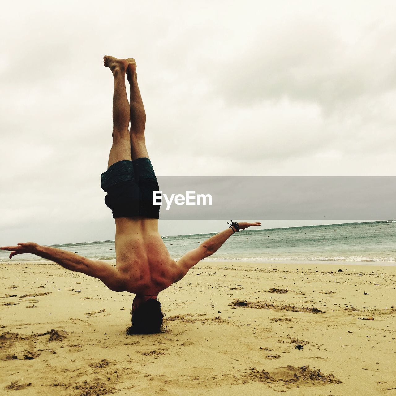 Shirtless man doing headstand at beach against cloudy sky