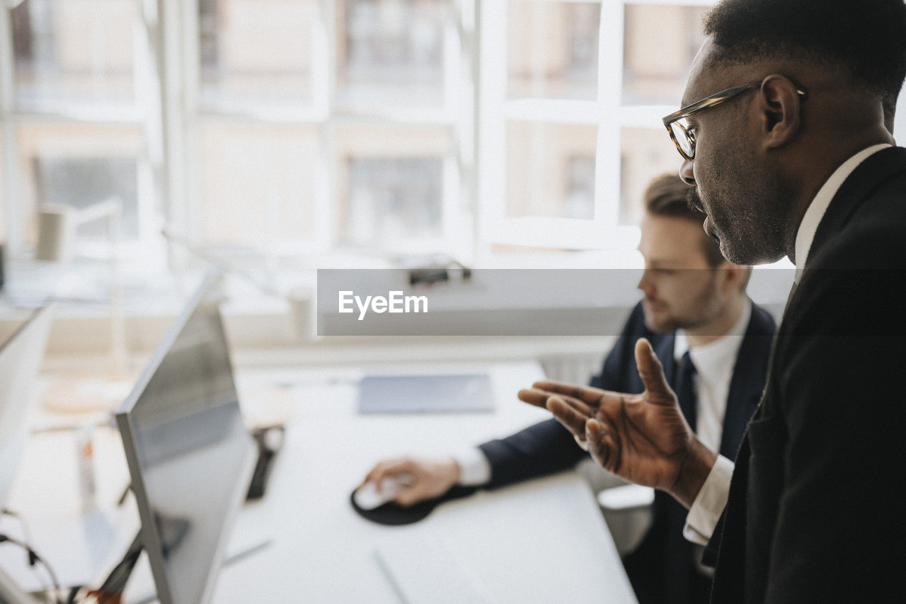 Businessman gesturing and explaining male entrepreneur over desktop pc at office