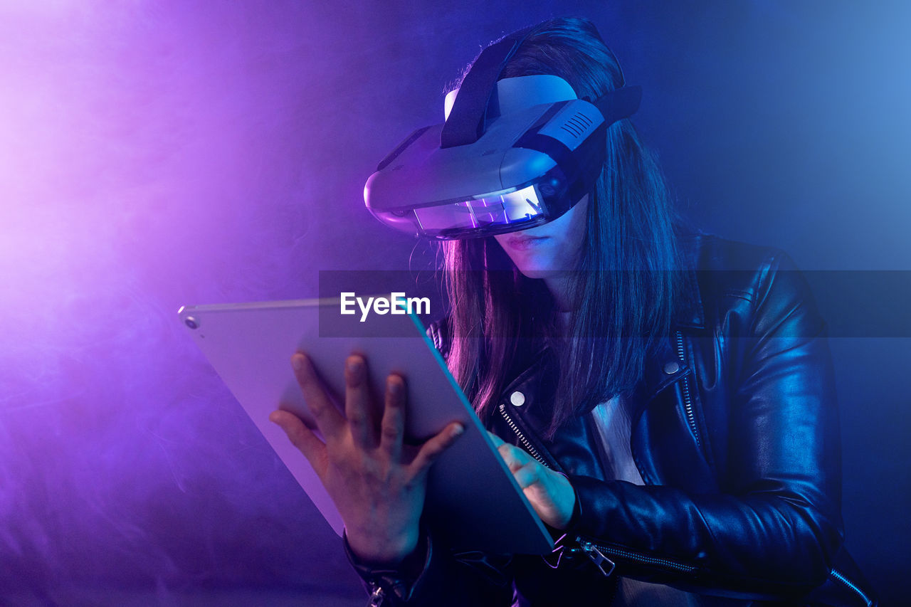 Concentrated anonymous female wearing modern vr headset standing near wall in dark room with contemporary tablet in hands under neon illumination