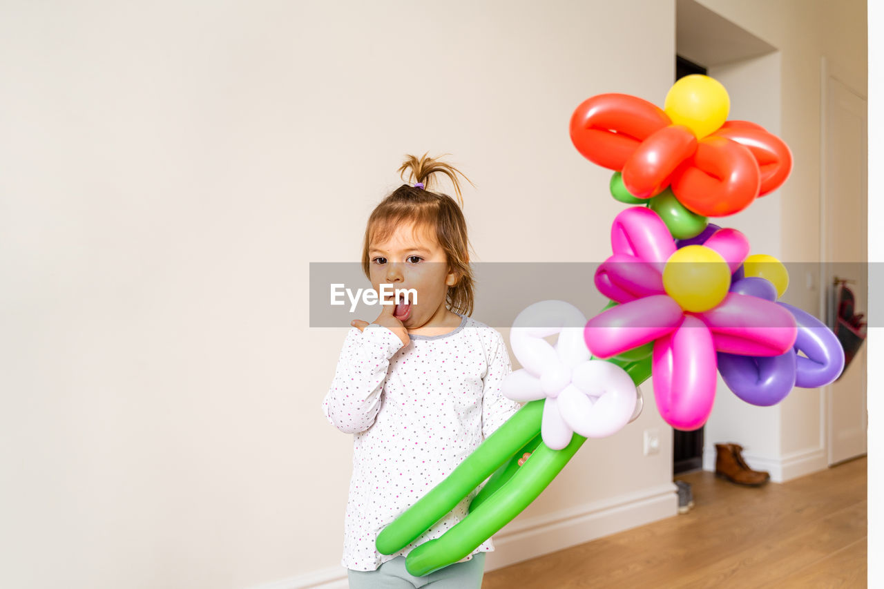 Cute girl with balloons standing against wall at home
