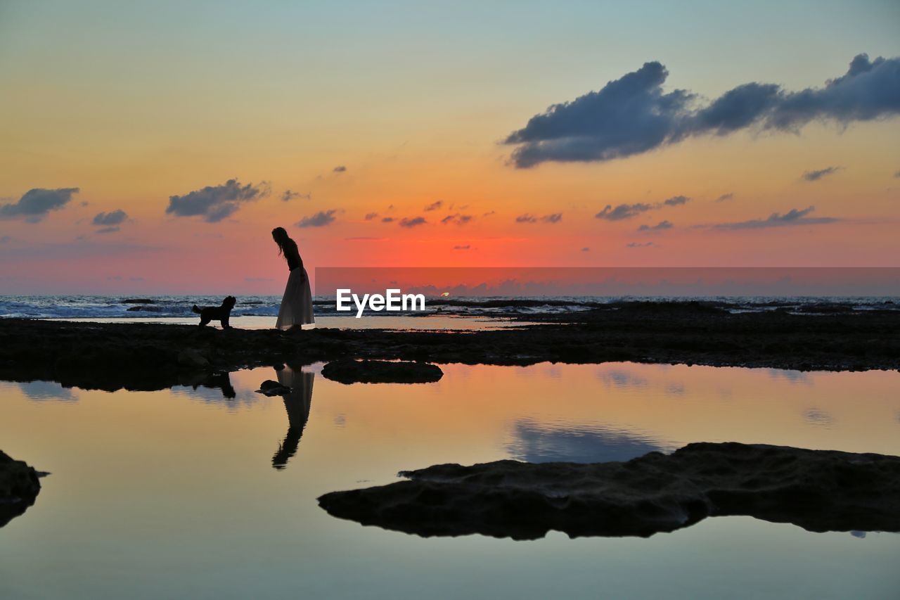 Silhouette woman with dog on rock at beach against sky during sunset