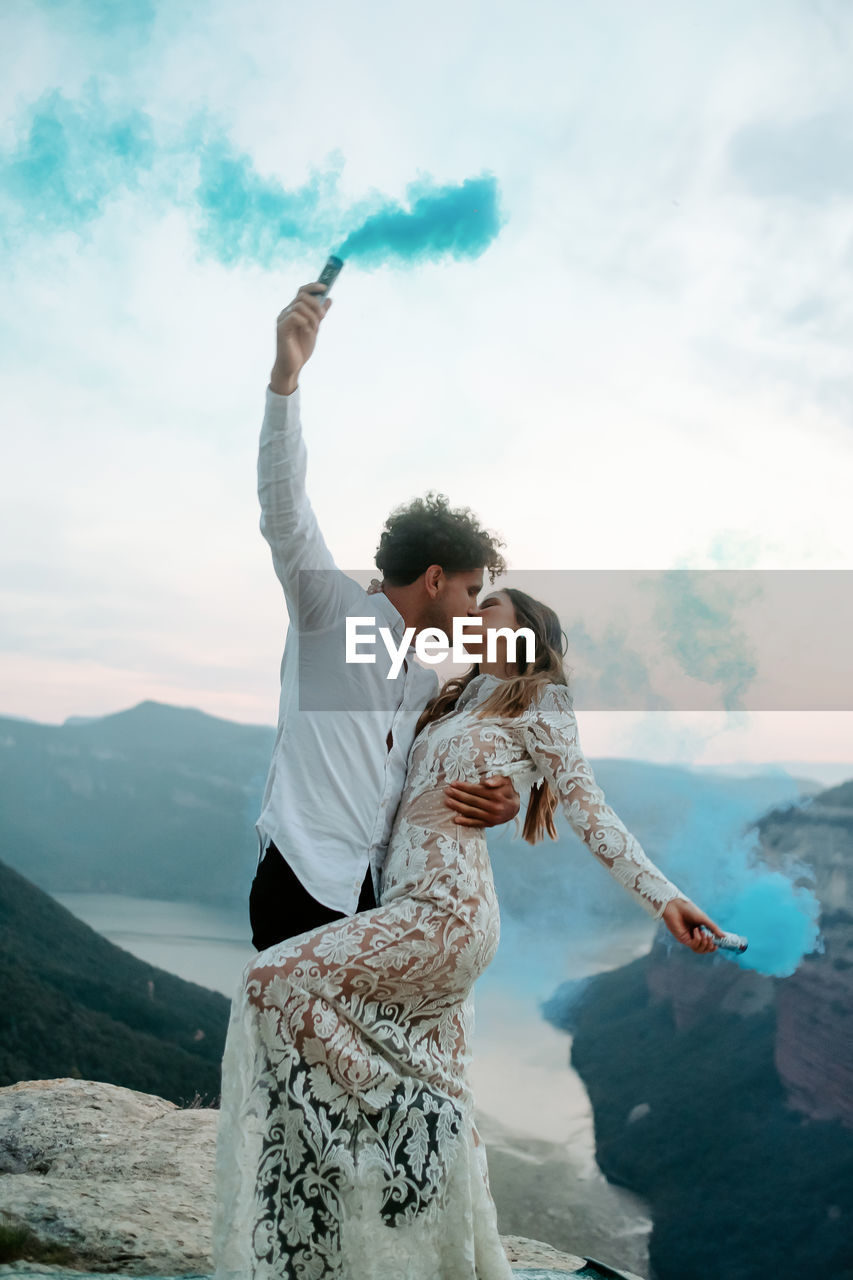 Full body side view of happy young newly married couple in elegant outfits holding colorful smoke bombs and kissing while celebrating marriage on top of rock in morro de labella in spain