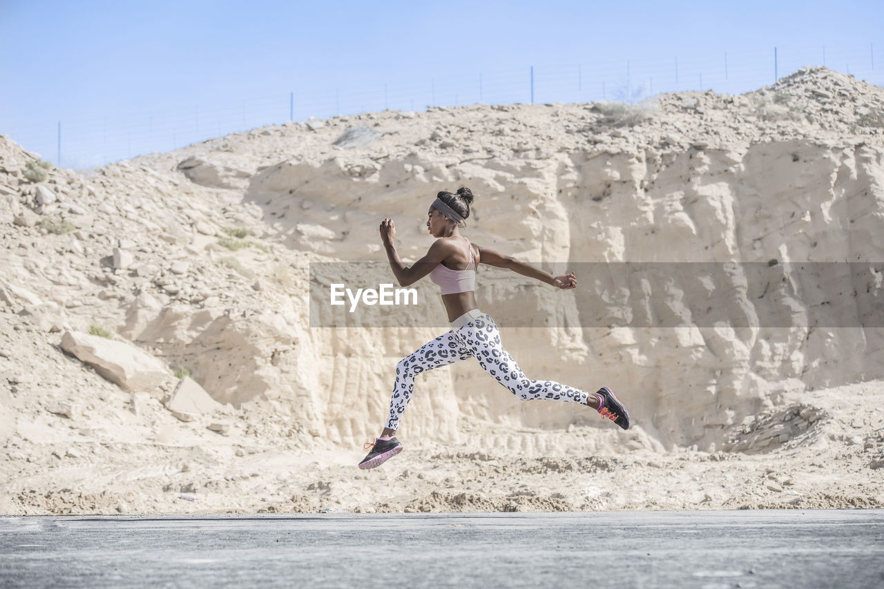 Side view of female athlete running on road against rock formation during sunny day