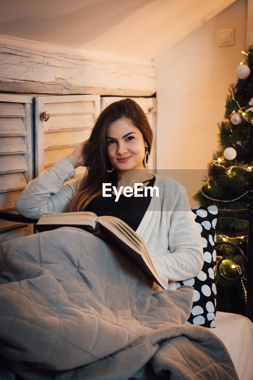Portrait of woman reading book while sitting on bed at home during christmas