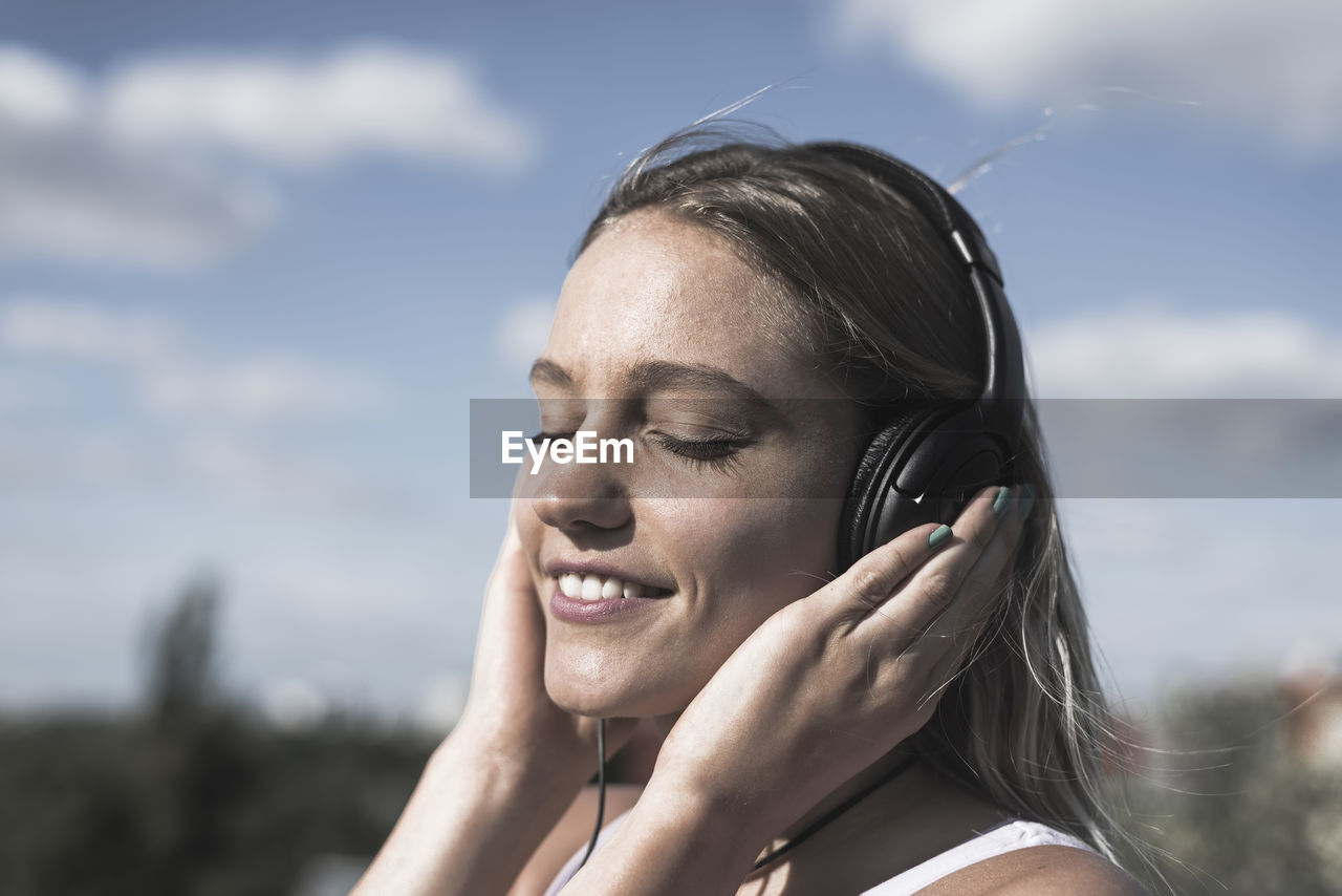 Close-up of young woman listening to music against sky