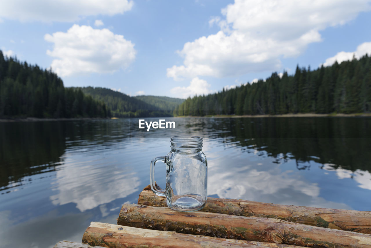 Jar on logs floating over lake against cloudy sky