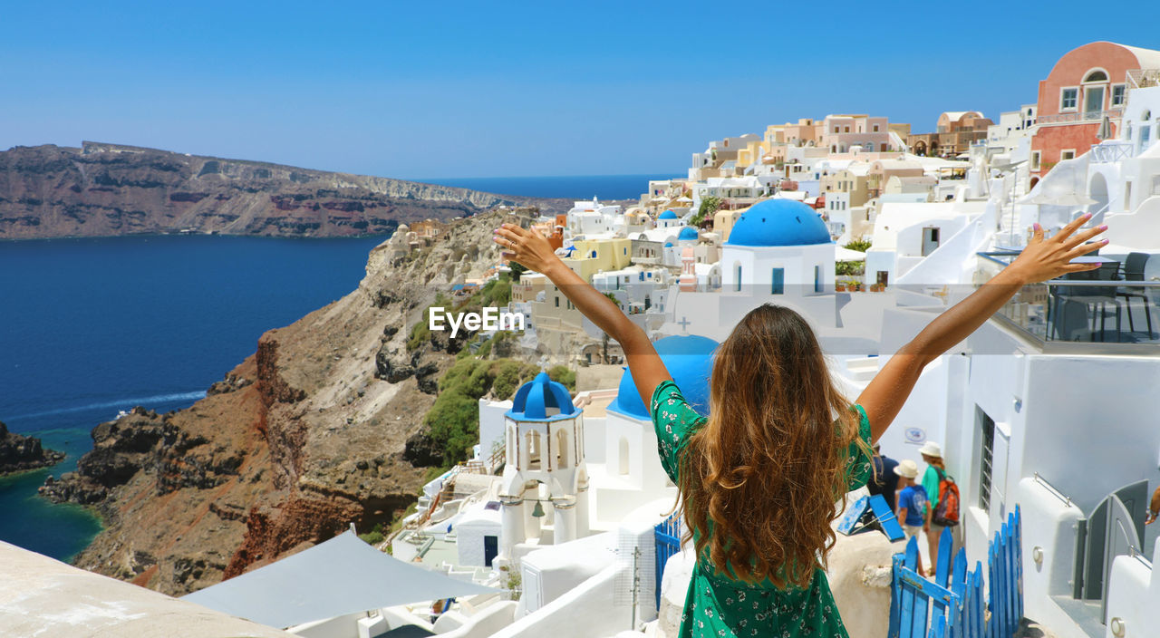 Rear view of woman with arms raised looking at houses against clear sky