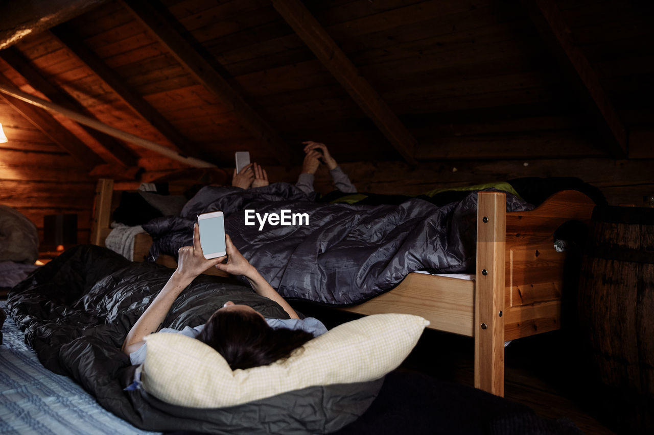 Male and female friends using mobile phones while lying in cottage during bedtime