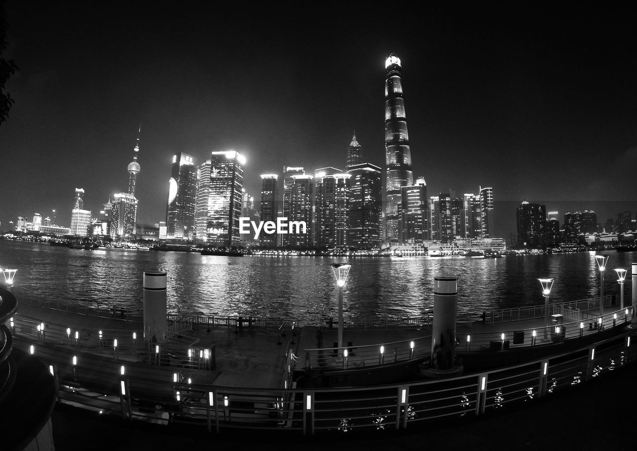 Illuminated modern buildings by river against sky at night