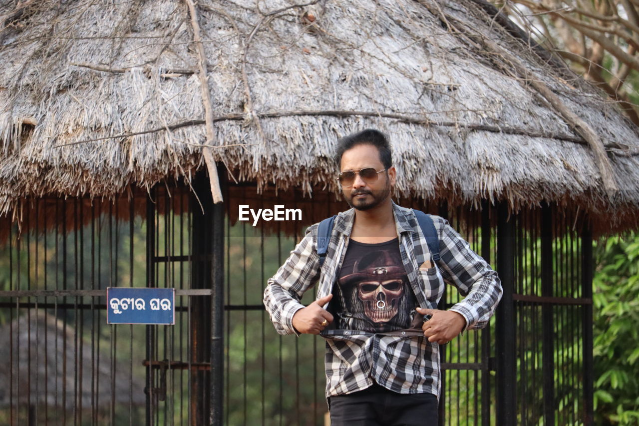 thatched roof, one person, adult, standing, portrait, hut, front view, men, young adult, roof, person, architecture, nature, thatching, looking at camera, spring, lifestyles, outdoors, clothing, day, three quarter length, holding, smiling, casual clothing, land, communication, leisure activity, tree, protection, rural scene