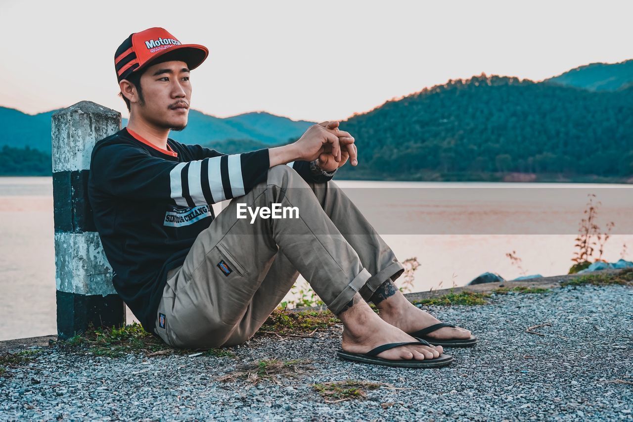 Young man looking away while sitting by lake