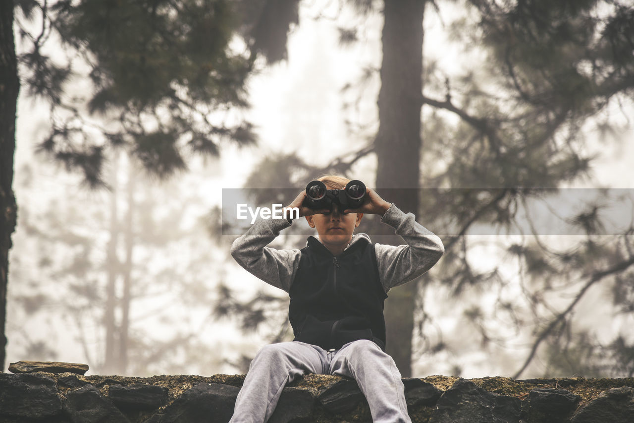 Boy looking through binoculars while sitting on retaining wall at forest