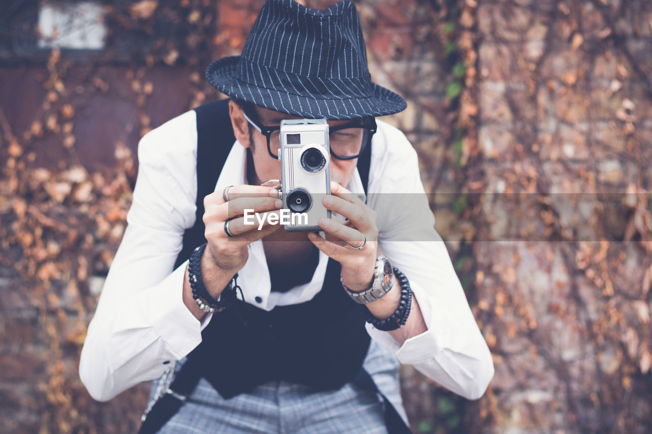 Mature man photographing while standing against brick wall during autumn