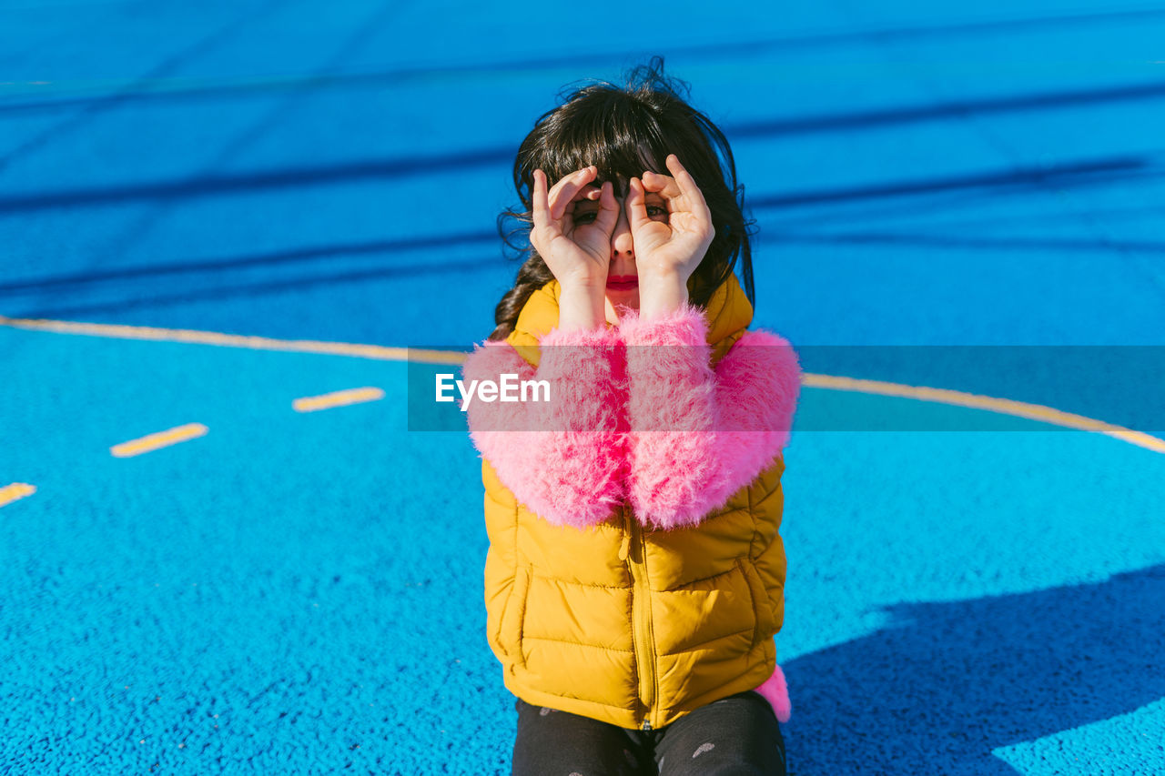 Girl forming binoculars with fingers while sitting at sports court