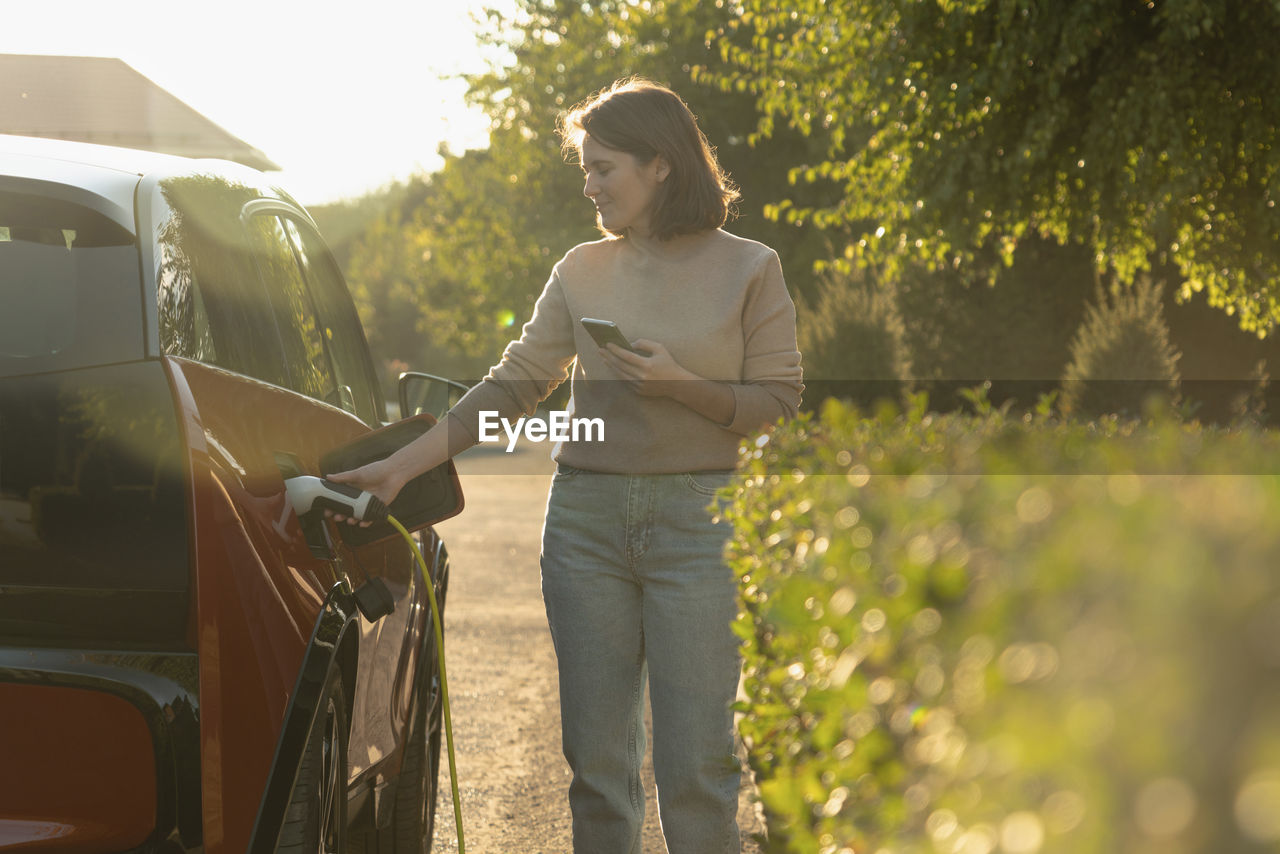 Woman with mobile phone charging electric car at roadside