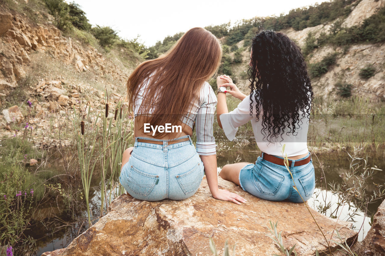 Two young attractive women best friends from behind sitting on stone end enjoying in nature 