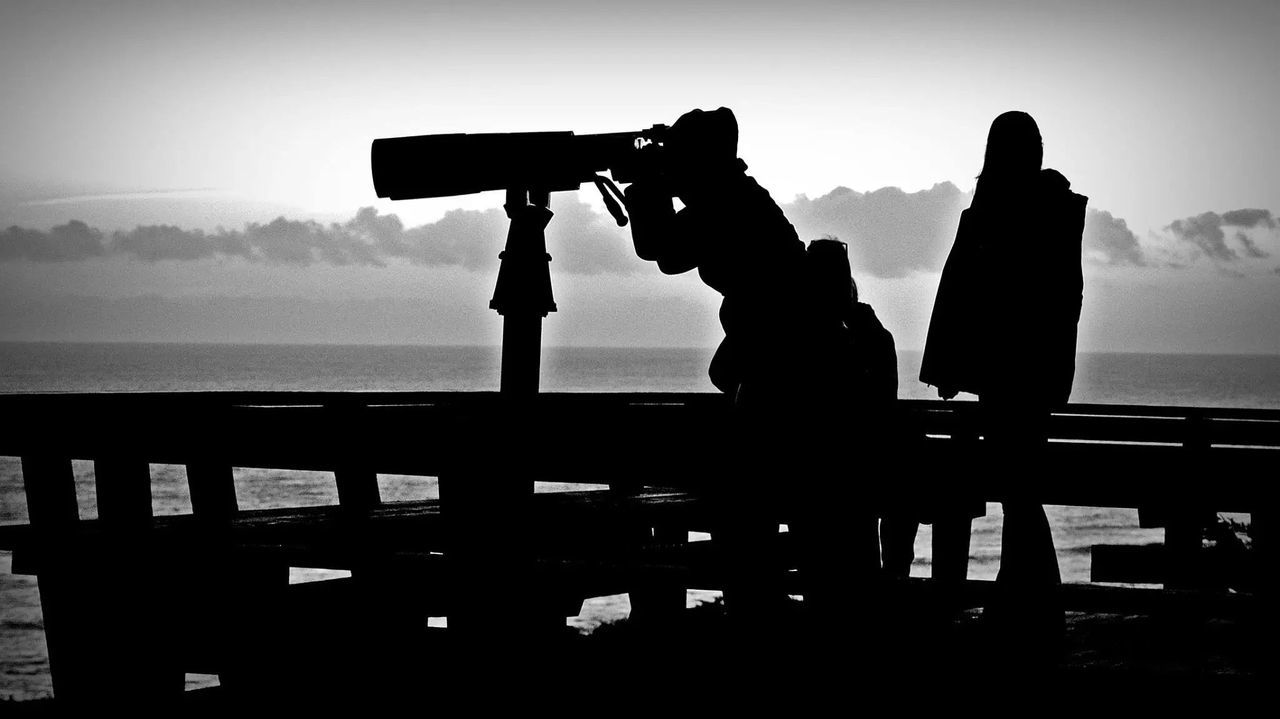 SILHOUETTE MAN PHOTOGRAPHING WOMAN WITH ARMS OUTSTRETCHED AGAINST SKY