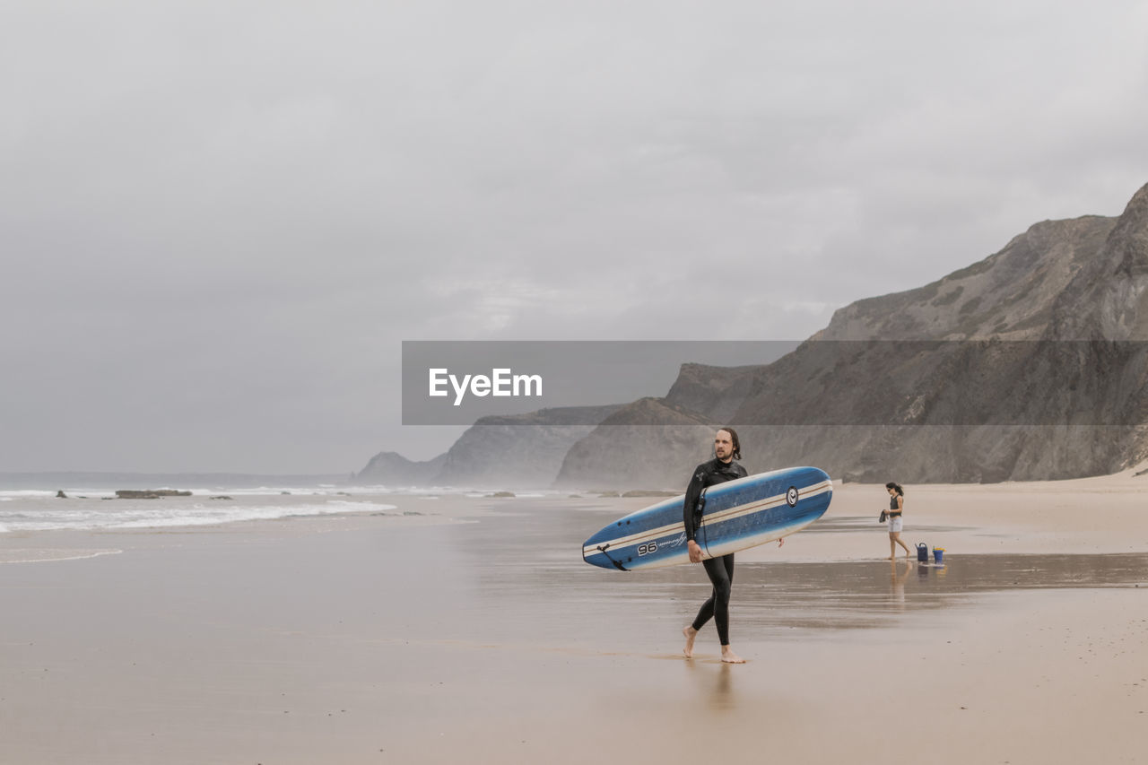 MAN STANDING ON BEACH AGAINST MOUNTAIN