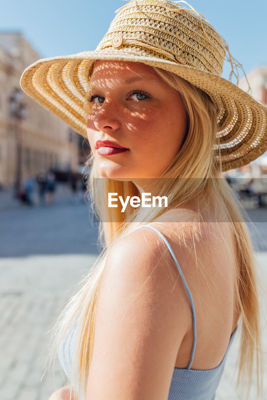 Side view of charming female wearing straw hat looking at camera on sunny day in city street in summer