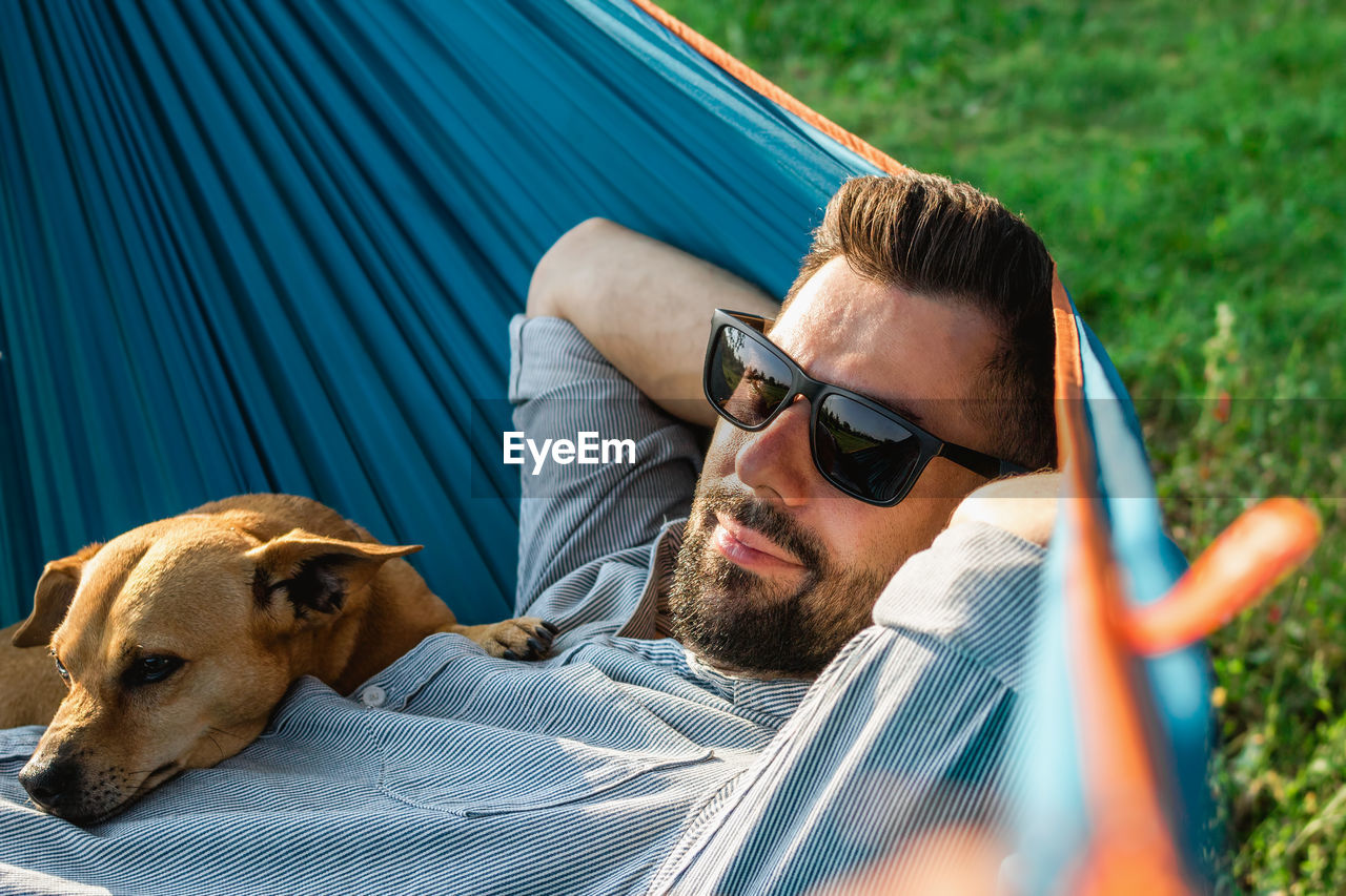 Young handsome european man in sunglasses is resting in hammock with his cute little dog.