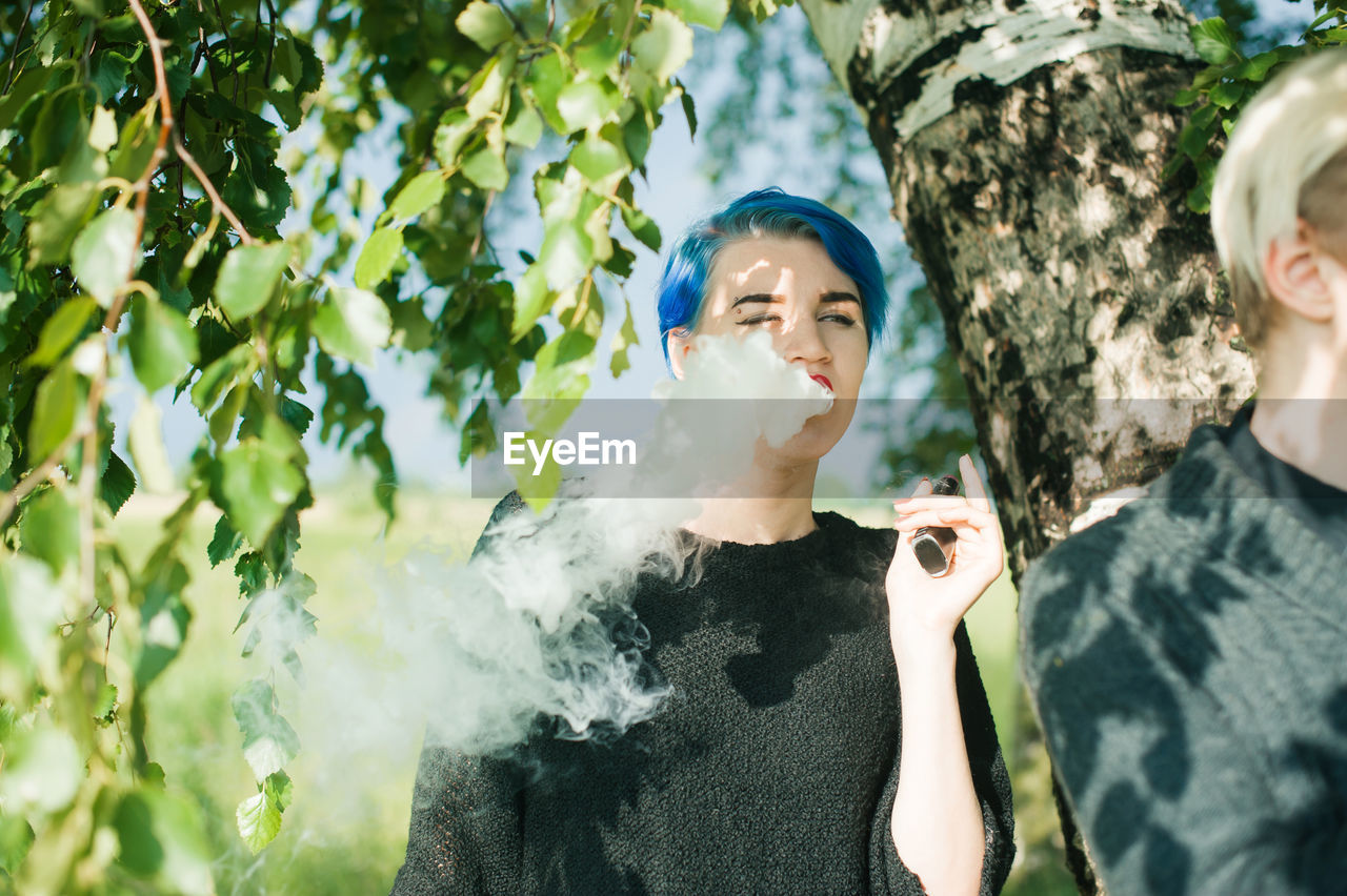 Young woman smoking electronic cigarette by tree