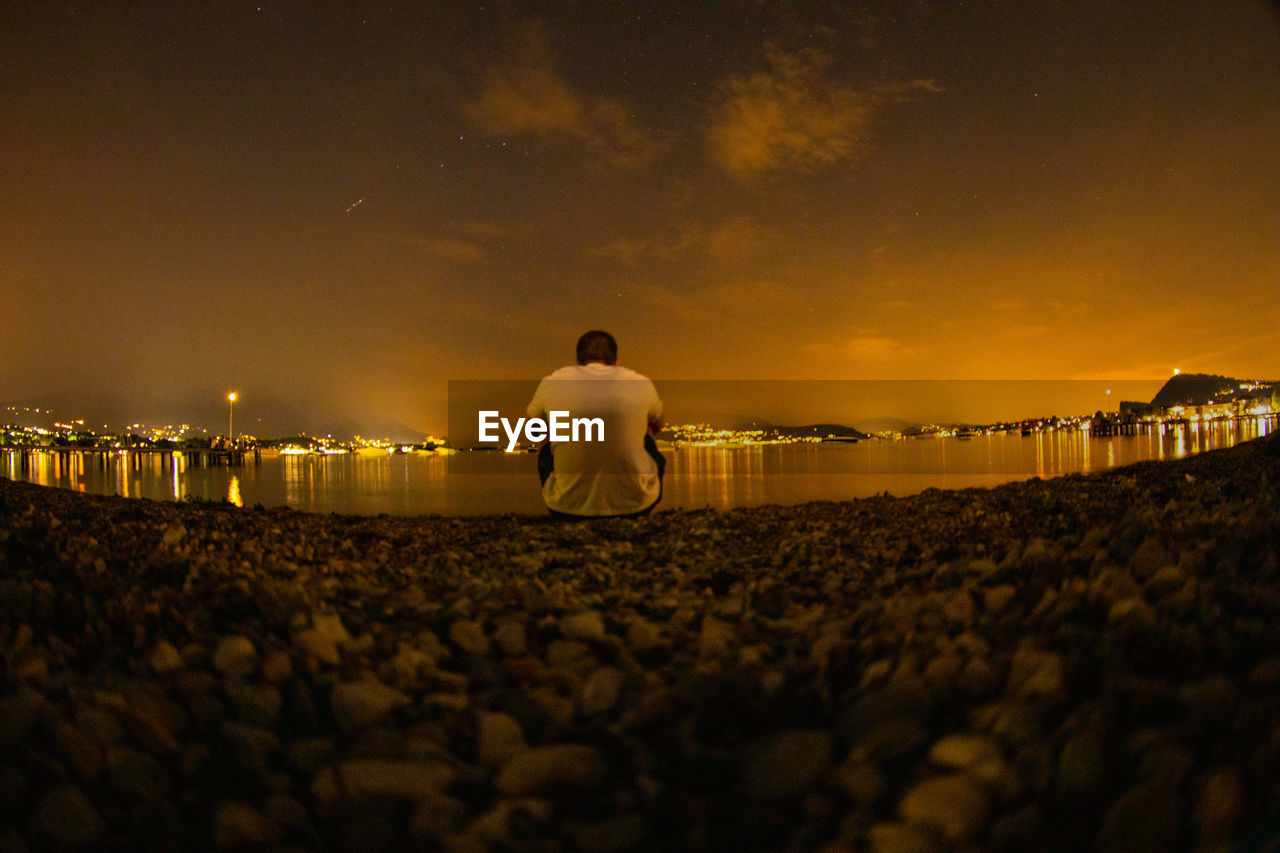 Rear view of man sitting at lakeshore during sunset