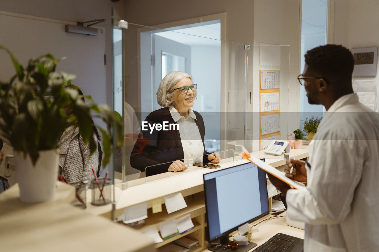 Smiling senior female patient communicating with male receptionist through transparent shield in clinic