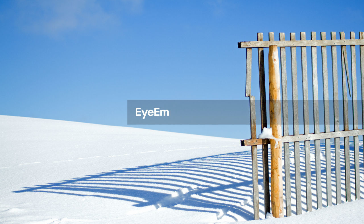 Shadow of fence on snow covered landscape against blue sky