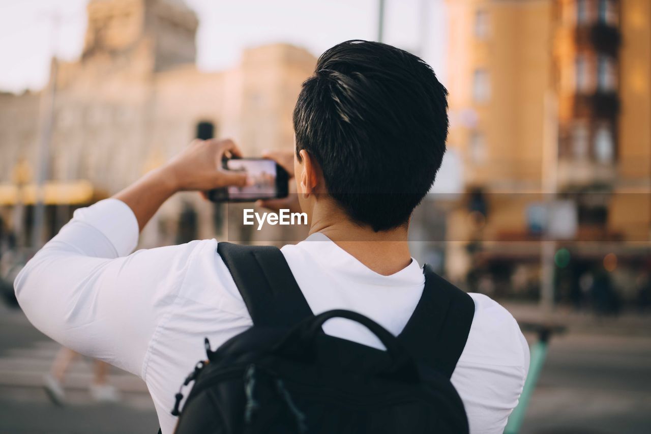 Rear view of young man photographing through mobile phone in city