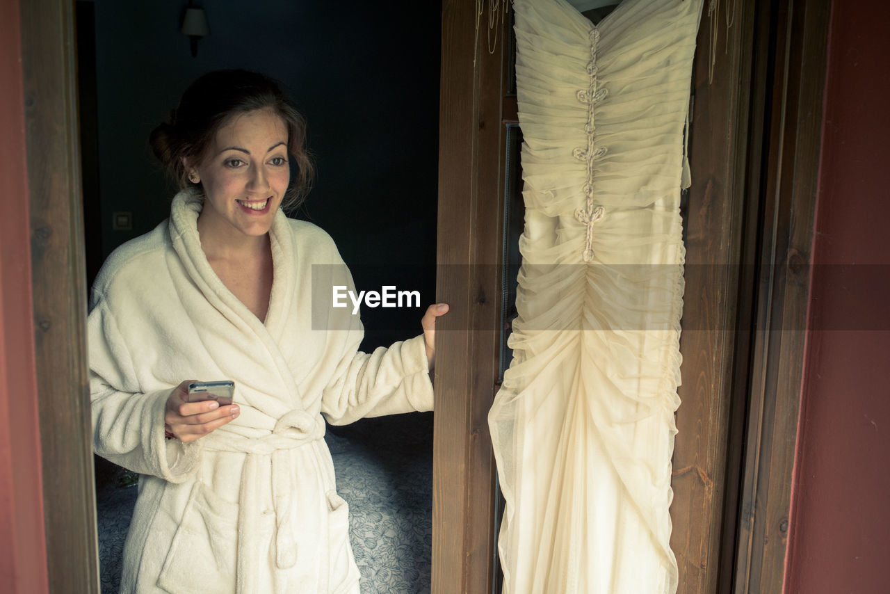 Smiling bride in bathrobe standing wedding dress on door in bedroom