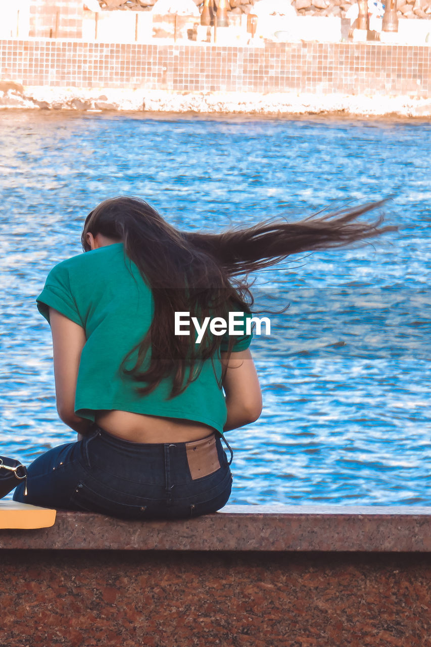 Rear view of woman sitting on ledge at riverside