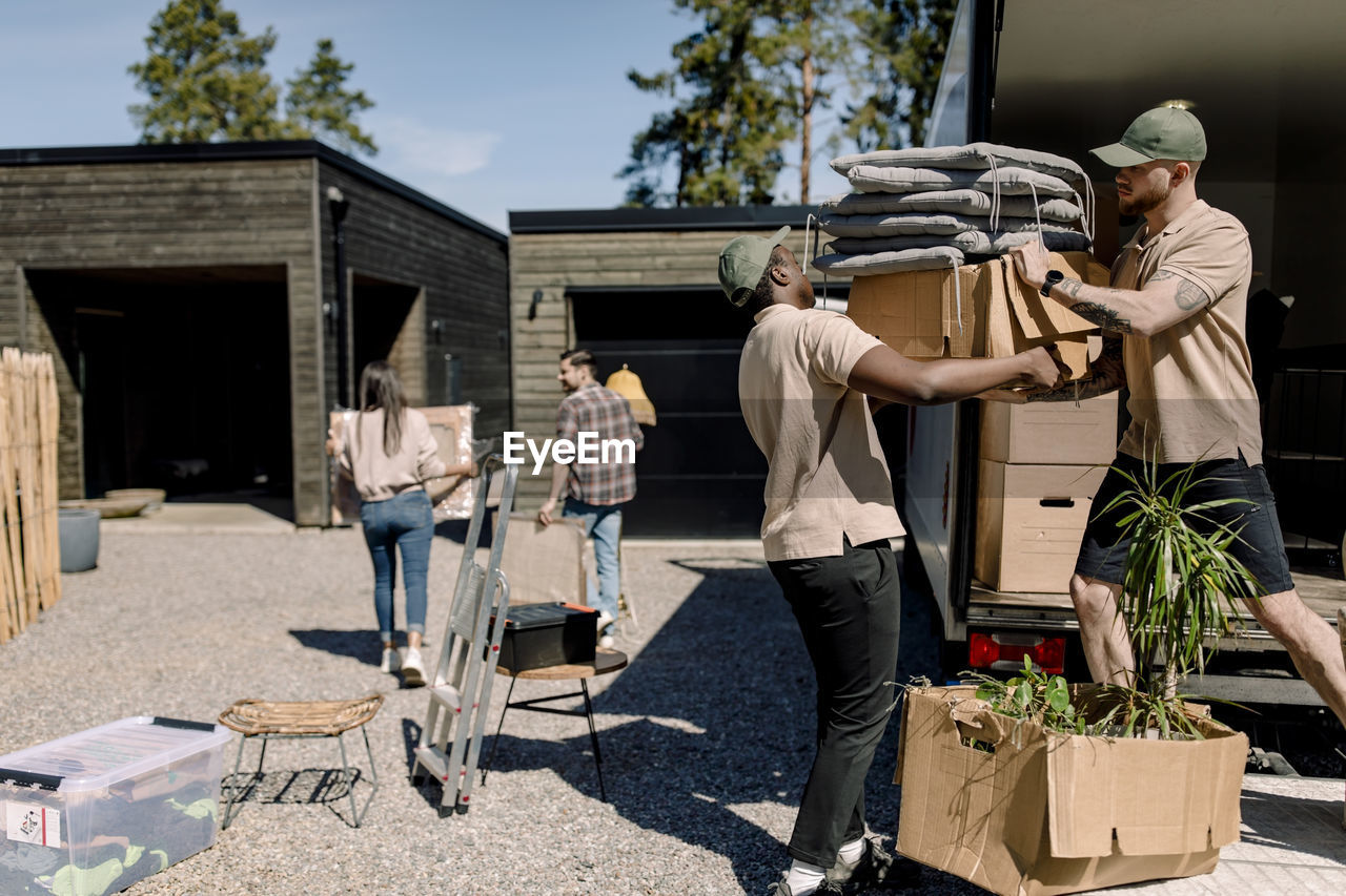 Multiracial movers unloading boxes from truck while couple in background