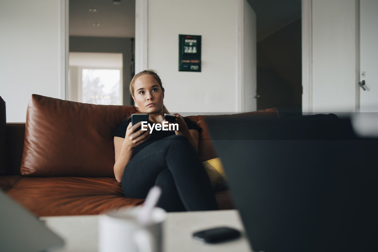 Teenage girl sitting on sofa using smart phone and remote control while watching tv at home