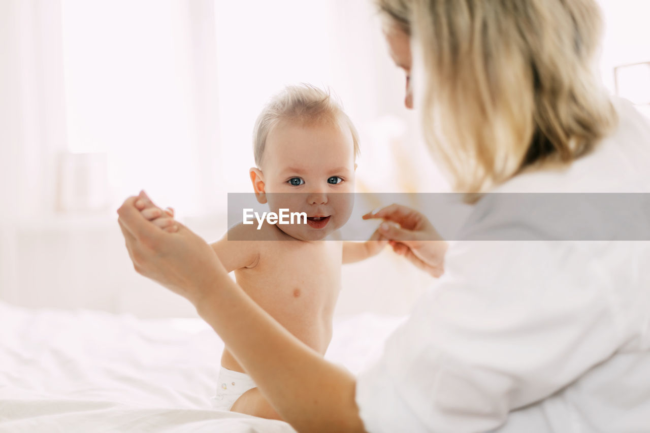 A cute baby is sitting on the bed with her arms outstretched and holding her mother's hands. 