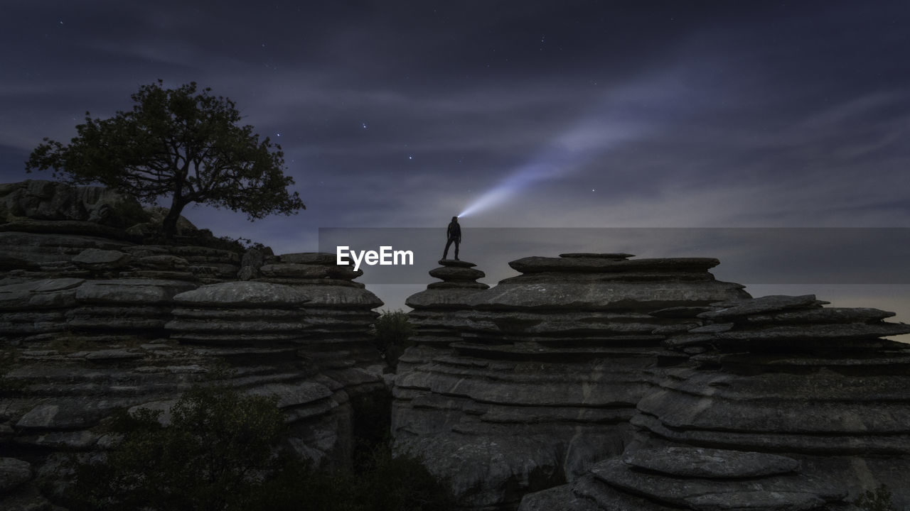 Silhouette man with illuminated flashlight standing on cliff against sky at night